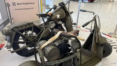A display with a barbed wire barrier. There a two motobikes, picture taken from the side of the motorbokes, a Harley-Davidson WLA and a Cushman Airborne Scooter in front.