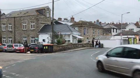 A silver car on a road bending to the left with car parks in bays on the left-hand side. There are stone buildings in the backgrlund