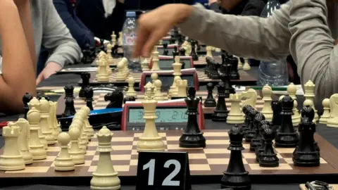 A row of chess boards being used for games. A hand is extending out from the right and preparing to make a move on one of the boards.