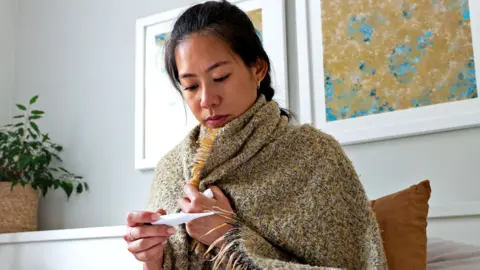 A young woman sits at home with a shawl wrapped around her looking at an oral thermometer