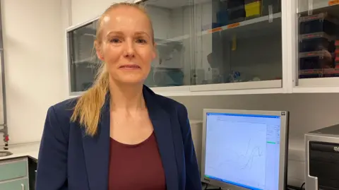 A woman with long blonde hair, wearing a blue jacket and red top. There is a monitor behind her.