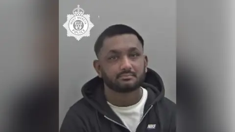 A police mugshot showing a man with very short dark hair with a dark beard wearing a white T-shirt and a black hoodie. 