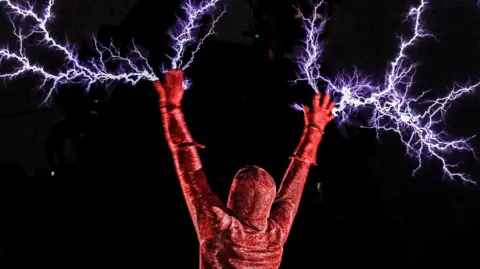 A person covered entirely by a red glittery body suit is pictured with their back to the camera with both arms raised in the air. Bright white-blue lighting bolts appear to burst from their hands against a background of total darkness. 
