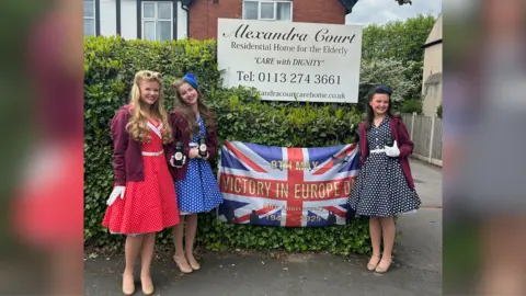 Duchy Darlings Siena, Emilia and Lottie stand outside Alexandra Court, a residential care home. All three girls have long hair worn down with 1940s-style victory rolls. They wear blue 1940s tea polka-dot dresses.