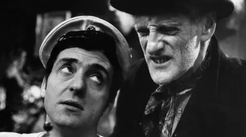 A black and white image of a young man staring up at an older man. The younger man has a white hat and the older man is wearing a neck scarf and black jacket.