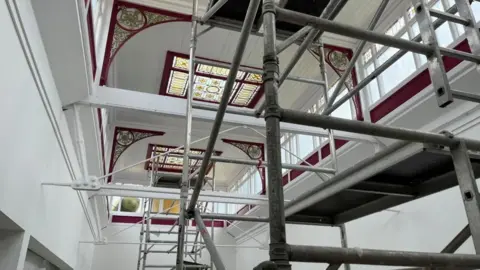 Andrew Turner/BBC Cast iron brackets and timber, painted in white and deep plum colour, and stained glass windows form a lantern in the former department store. Scaffolding towers have been erected in the near ground and background of the image.