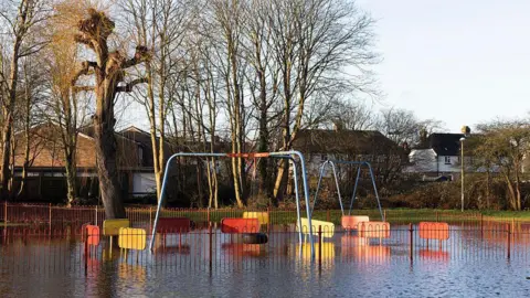 Getty Images Two sets of swings stand in a flooded playground surrounded by railings, with trees and houses visible in the background