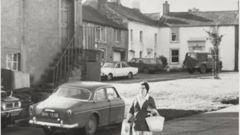 The Hesket Newmarket Community Shop Black and white picture of woman in the 1960s walking through the village in front of the shop while carrying shopping bags