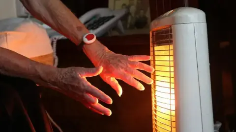 PA Media An elderly person is once heating his hands with fire. The fire is in a white plastic case and the front is a silver grill at the front. There is an orange glow from inside the grill. Only the hands of the person are visible and both of them are holding the heater's body.