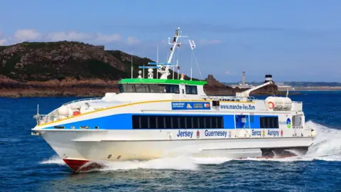 Manche Iles Express A ferry at sea leaving a wake behind it on a blue sky day. Lettering on the boat reads Jersey, Guernsey, Serq, Aurigny and a web address.com. There is land to its starboard side.