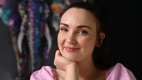 Diona Doherty, with brown hair, tied back. She is wearing a pink top and is holding one hand up to her chin. 