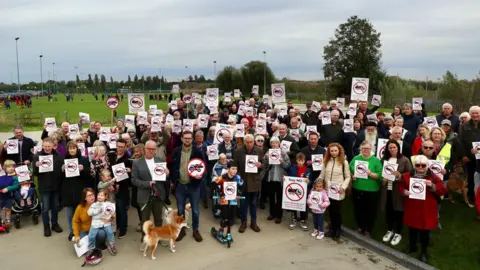 Protesters with placards about the proposed concrete crushing facility at Moorend Lane in Thame. They are looking at the camera. Among the adults, there are children and dogs as well.
