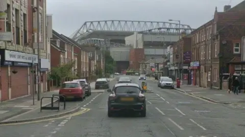 Google A wide single carriage road leads to Anfield stadium. There are a number of parked cars in front of shops.