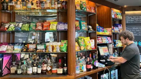 BBC A bartender behind a pub bar with many crisps on show