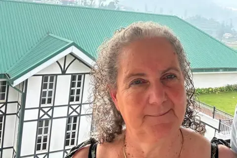 Alan Dutton A woman in her 60s with grey curly hair pulled back from her face stands in front and slightly above of a large white building with black window frames and a green roof. She is wearing and has tie straps on her shoulders. In the distance a misty hill side is in view.