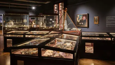 Cornwall Museum & Art Gallery The photo shows an interior view of a museum exhibit. The room contains multiple glass display cases showcasing various rock specimens. The display cases are illuminated. There are more display cases in the background with some paintings on the wall. The room has wooden flooring and dark-coloured walls.