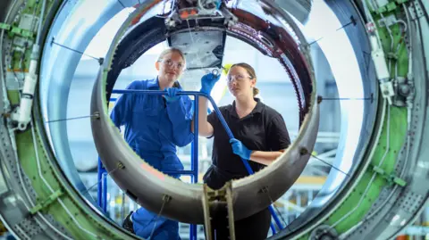 Getty Images A female apprentice, in blue overalls, is shown jet engine housing by an instructor, wearing black overalls - they are both wearing safety goggles and blue gloves 