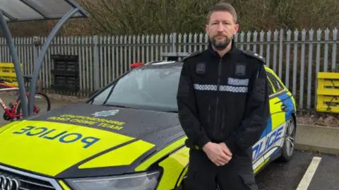 Staffordshire Police A police officer, in a black police jacket and trousers is standing to the side of a liveried police estate car in a compound.