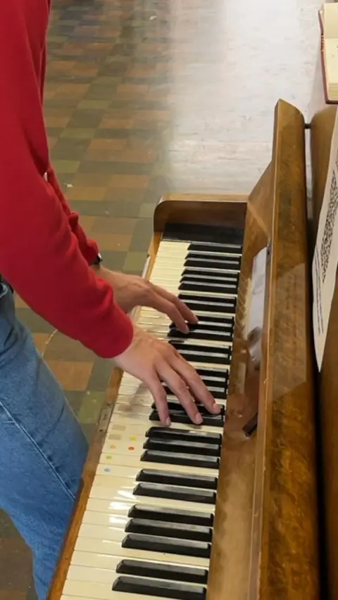 A close-up of piano keys. A man's hands and arms are also in the picture. They are wearing a red top and jeans. 