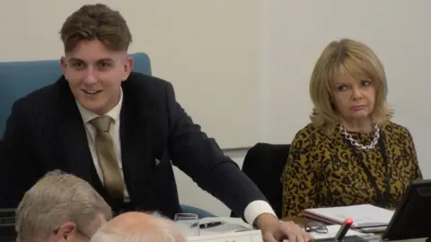 George Finch and Monica Fogarty sitting at a table during a meeting. Finch has short brown hair and is wearing a suit. He has his left hand on the table and is looking to his right while smiling. Fogarty is wearing a silver necklace and a yellow and black top. She is tilting her head to the right while looking towards Finch.