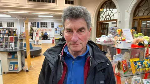 BBC/RICHARD EDWARDS A man with short dark hair and is wearing a blue fleece and a black jacket. Books and soft toys are on a shelf behind him.