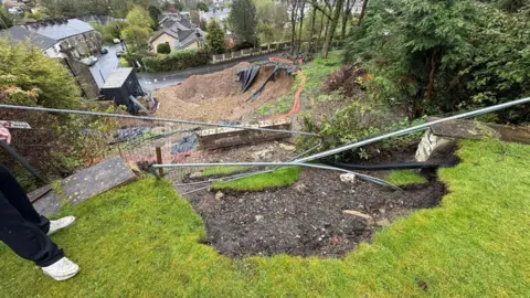 Bethanie Bailey A fence loosely hangs across a lawn on a steep garden that has fallen away into a large pit, littered with construction materials. A person's feet and legs can be seen to one side. 