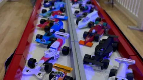 Miniature cars on a model racetrack. The cars are sat diagonally across the track. The track is on a wooden floor. 