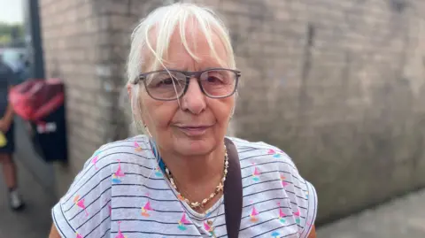 Gillian Sims in grey t-shirt with pink print wearing a shell necklace and glasses and grey hair tied back
