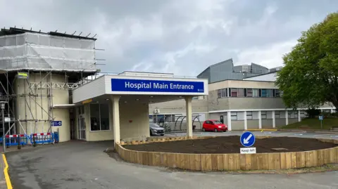 A view of Guernsey's Elizabeth Hospital showing the main entrance. It shows a single story building with scaffolding and cars. 