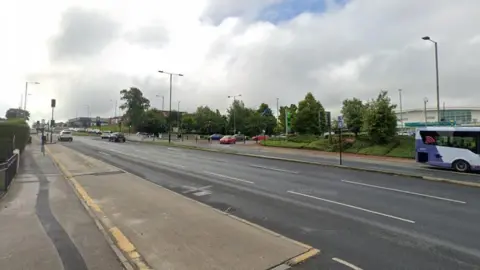 A street view image of the road shows several cars on either side. Trees line it in the background.