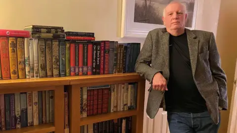Mark Wood Keith Wood is standing with his right arm leaning on a bookcase. He is wearing a brown jacket, black top and blue jeans.