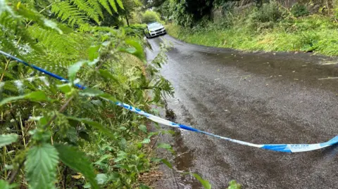 Police tape lying across a narrow country lane. There is green vegetation on either side of the road, and a police car is visible at the far end . 