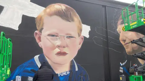 A giant mural of musician and Ipswich Town fan Ed Sheeran is pictured halfway through being created in the Suffolk town. On the left is a picture of Sheeran as a boy in an older Ipswich Town kit, and on the right he is seen as he looks now. A green cherry picker crane is visible and also a man in a black hooded coat who is working on the mural