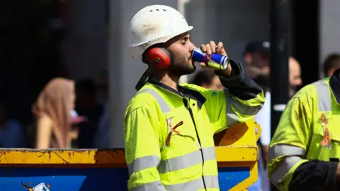 Reuters A sweating workman wearing a helmet and hi-vis protective clothing takes a sip from a cold canned drink. 