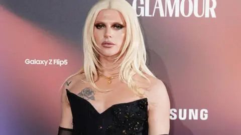 Bimini Bon Boulash, with blonde midlength hair and wearing a black sequinned corset, stands against a pink and grey promotional background.