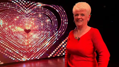 Sheila Diamond is standing on stage in front of a screen with a sparkly heart. She is wearing a red, long-sleeved top and is smiling at the camera. 