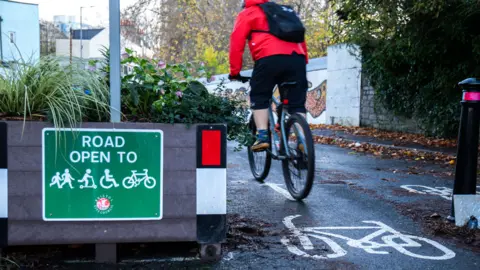 Bristol City Council A cyclist seen from behind wearing a red jacket and black backpack rides past a roadside planter. A green sign on the planter reads “Road Open To” with symbols for pedestrians, wheelchair users, scooter users and cyclists, alongside a Bristol City Council logo.