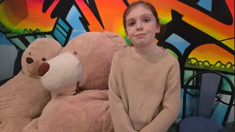 A young girl with ginger hair and freckles wearing a salmon-coloured jumper is sitting next to two large teddy bears which look about the same size as her. She is in front of a bright orange and blue mural on a wall and is smiling at the camera. 