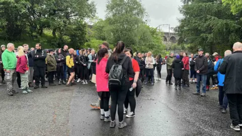 a large gathering of people in boots jackets and wet weather clothing gathers on tarmac near some trees