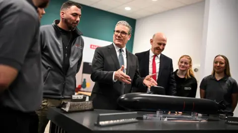 PA Media The prime minister and defence secretary with a small group of BAE staff in a large room. On a table in front of them is a model of a black submarine. Starmer is talking to the group and has his hands out towards the model.