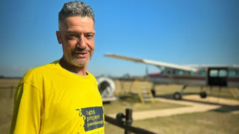 Emma Baugh/BBC Dan Perkins wearing a yellow T-shirt standing in front of a plane