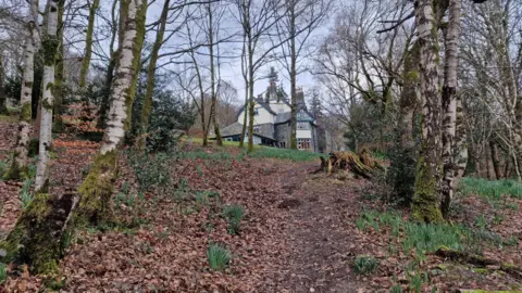 Hawes End. The Victorian building is made up of various sections, with numerous chimney pots on the pitched roof. It sits on top of a slope, which is covered in leaves and surrounded by birch trees.