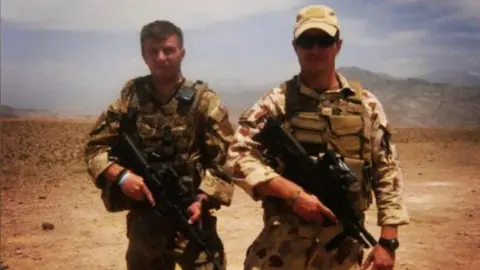 Lee Rackham Two men in camouflage gear, holding guns, standing in a desert. One has a cap and sunglasses on 