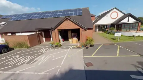 Google The entrance to Eden Valley Hospice is on a single site with a single story brick building on the left with a low row of solar panels. 