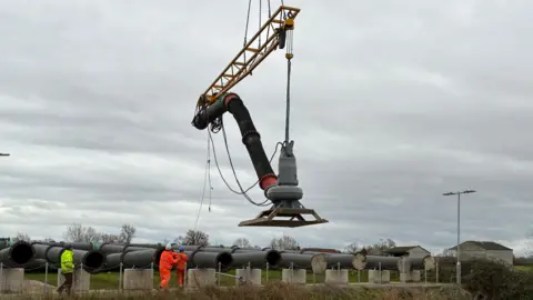 A crane lifts an industrial pump into a river in Somerset. There are three men in Hi Vis working below.