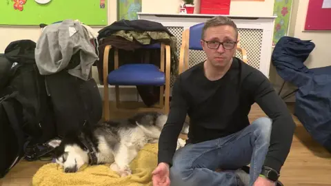 Lukasz sits on the floor wearing jeans, a black long-sleeved top and glasses. Next to him is his dog, a husky, who lies on a yellow blanket. 