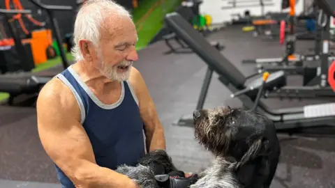 Ross Starkie Mick Wadge - a white-haired man in a vest top, surrounded by gym equipment - smiles at a black and grey dog standing on its hind legs 