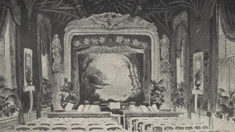 Wellcome Collection Black and white photo of the Gaiety Theatre at Plas Newydd, Anglesey. There are rows of seats in lines of five and six in front of an orchestra pit and a proscenium arch stage with curtains and a painted front curtain showing an island and seascape. The room has vaulted arches on the ceiling and lots of palm plants dotted around the edges.