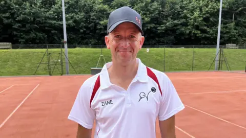 Leanne Rinne/BBC A man wearing a baseball hat and white tennis polo shirt standing on a tennis court 