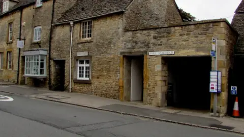 Stow-on-the-Wold town centre public toilets. They are based in a Cotswold stone building, and a bus stop can be seen to the left.